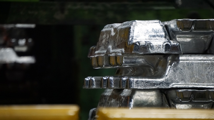 Several cast metal ingots are stacked on top of each other inside an industrial plant. The shiny silver surfaces catch the light, highlighting their rough texture and molded edges. The background is out of focus, showing dark machinery and green industrial structures, with yellow safety barriers partially visible in the foreground.