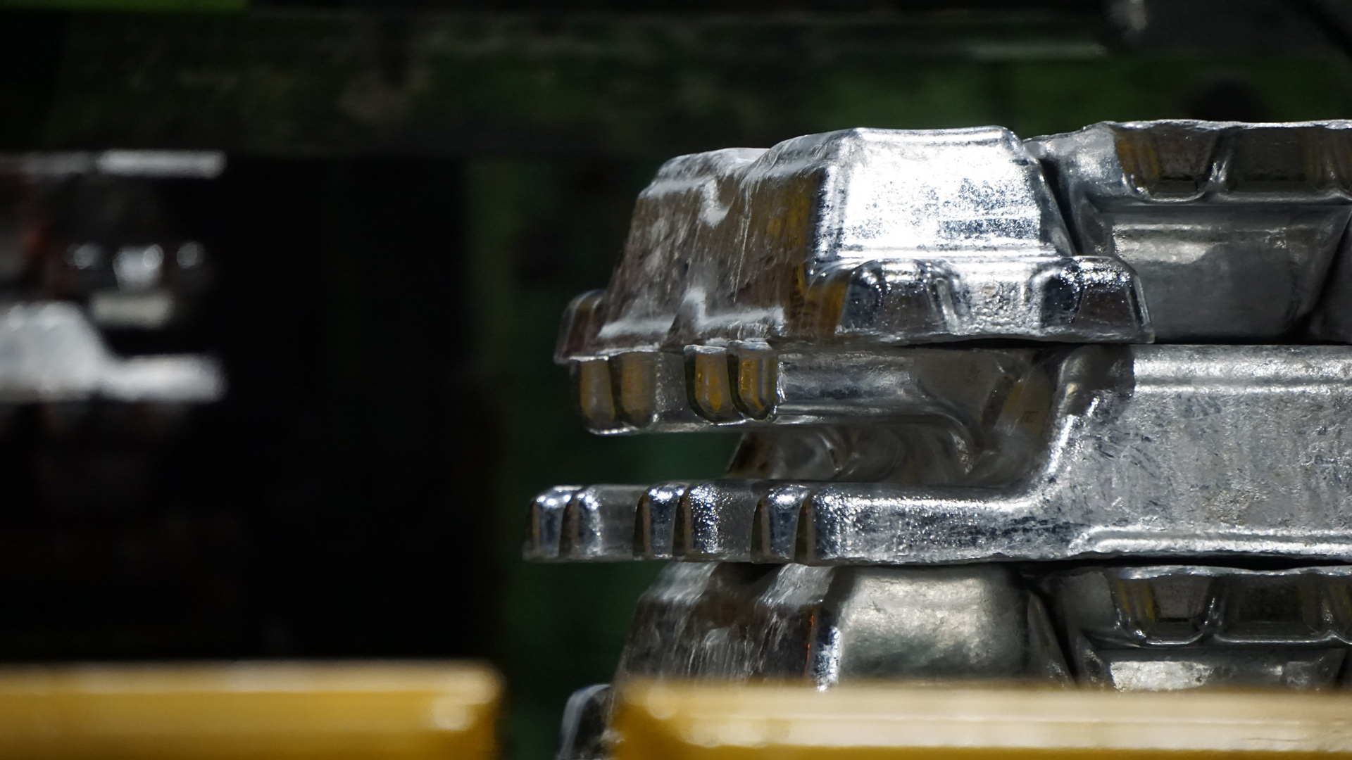 Several cast metal ingots are stacked on top of each other inside an industrial plant. The shiny silver surfaces catch the light, highlighting their rough texture and molded edges. The background is out of focus, showing dark machinery and green industrial structures, with yellow safety barriers partially visible in the foreground.