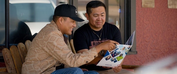 Two young men sitting on a chair, both looking at a brochure and discussing its content.