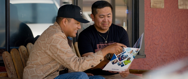 Two young men sitting on a chair, both looking at a brochure and discussing its content.