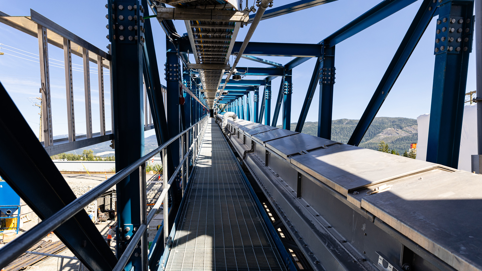 Covered conveyor system transporting bauxite ore through a steel-framed industrial structure, with mountainous terrain visible in the background.