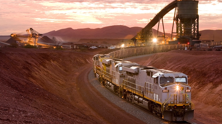 Freight train with multiple locomotives traveling along a curved track through an open iron ore mine at sunset, with conveyor belts, heavy machinery, and piles of ore illuminated by warm lights against a mountainous backdrop.