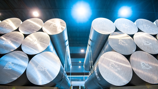 Stacked aluminium billets in a factory, illuminated by bright overhead lights.