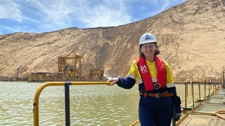 Person wearing a white hard hat, safety glasses, high‑visibility yellow shirt, gloves, and a red life jacket standing on a metal walkway beside greenish water at an industrial dredging or mining site. Yellow railings line the platform, and heavy machinery sits across the water, with steep sandy cliffs rising in the background under a partly cloudy sky.