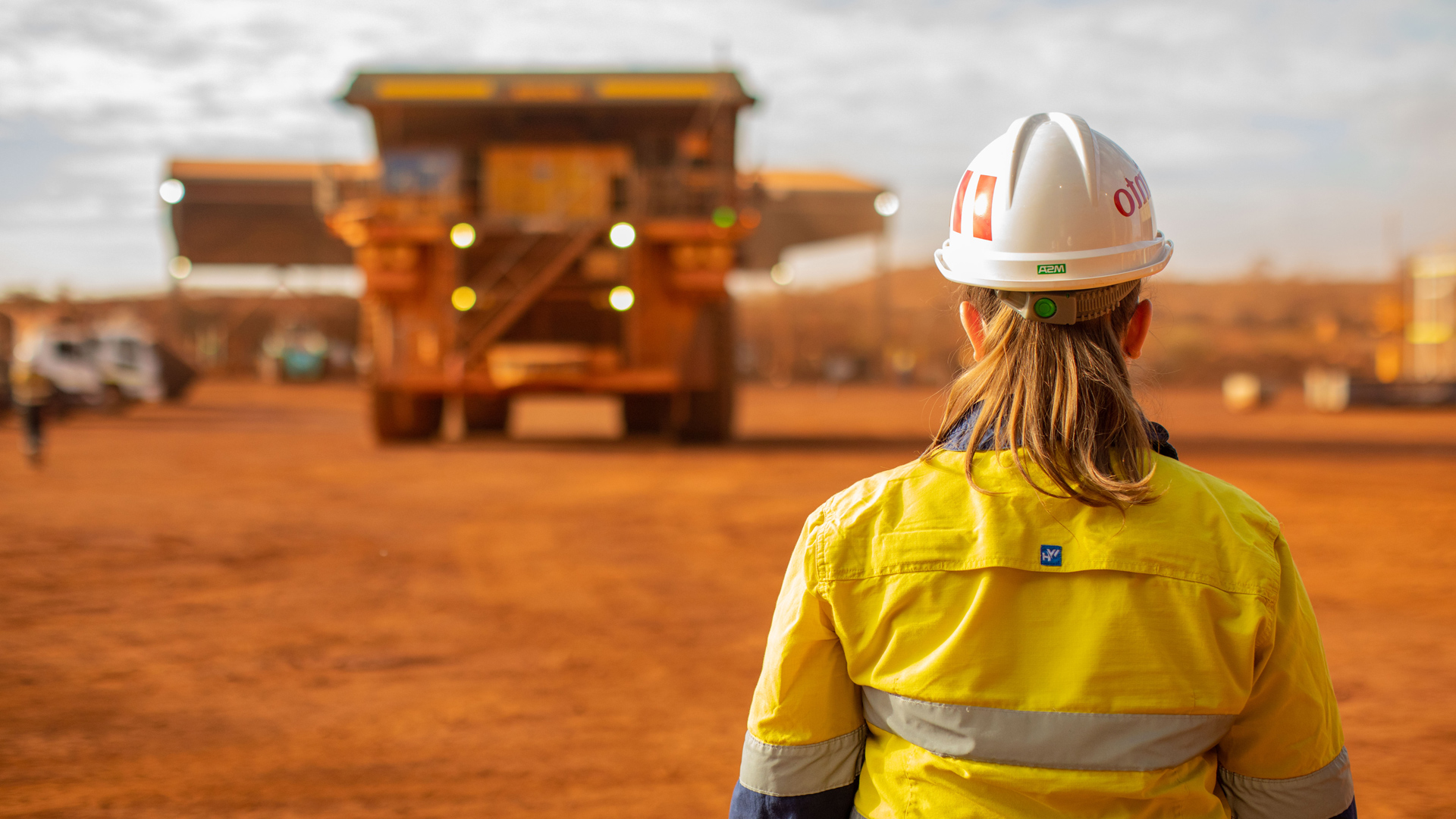 Person in a high-visibility yellow work shirt and white hard hat stands with their back to the camera, looking across a red dirt mining site toward a large haul truck in the distance under a partly cloudy sky.