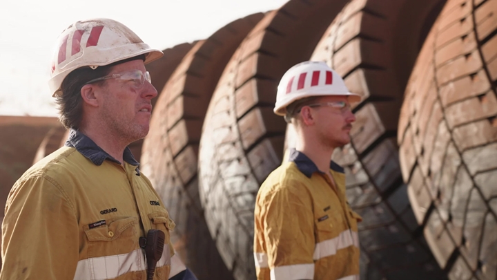 Two Rio Tinto employees wearing a safety hat and facing ahead in front of large mine truck tyres. 
