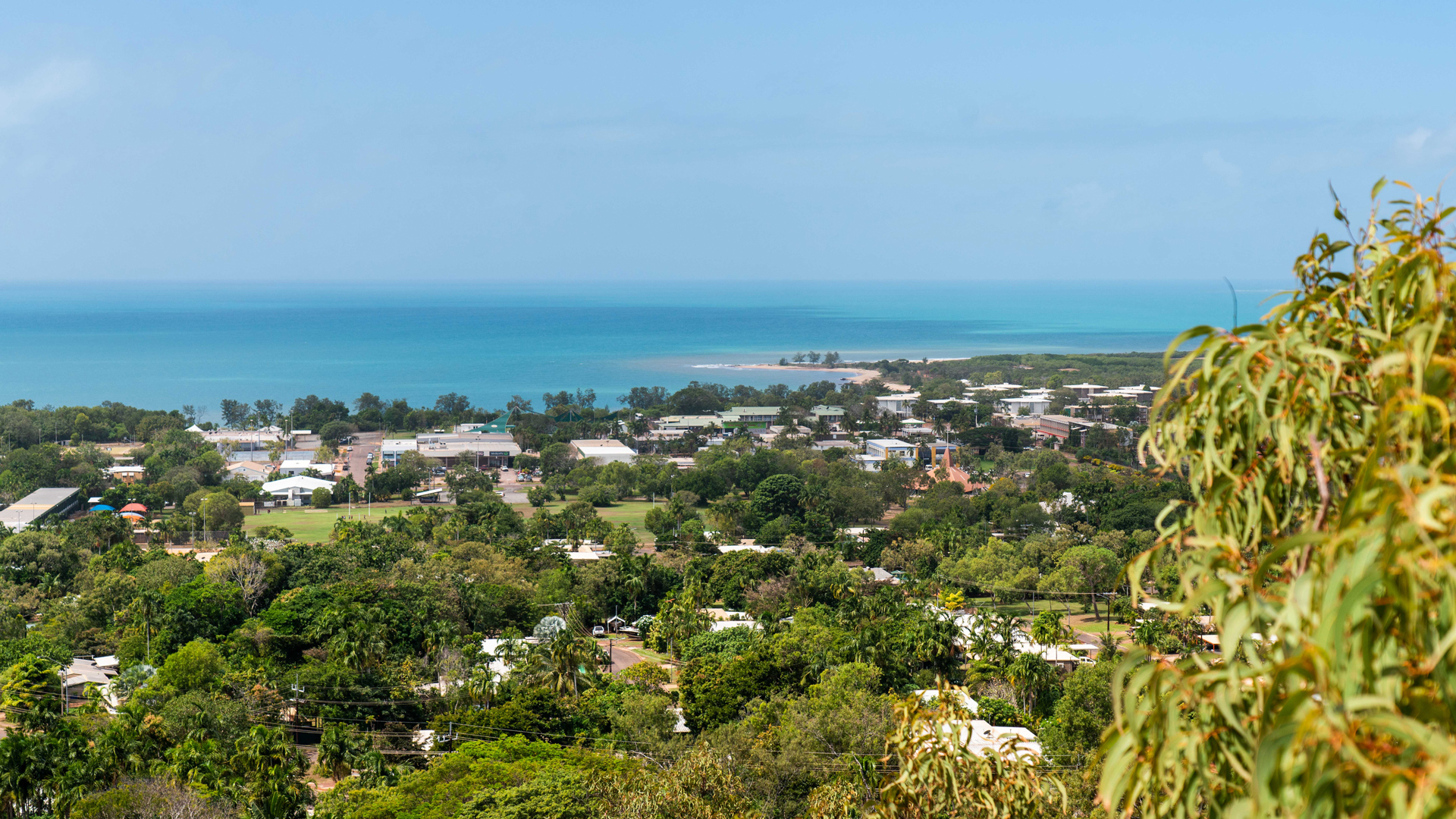 Aerial-style view of a tropical coastal settlement bordered by lush vegetation, featuring buildings, open green spaces, and a turquoise shoreline extending into the ocean.