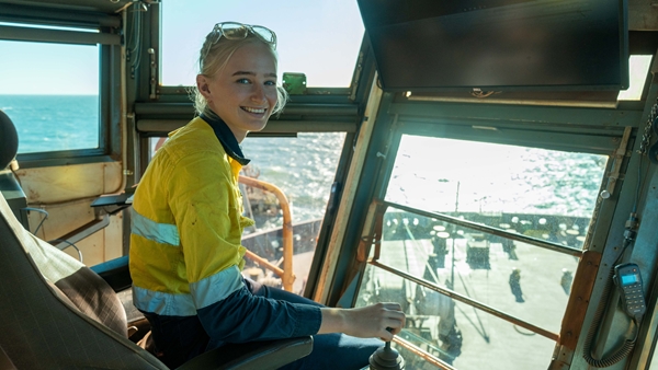 A worker in a high-visibility yellow jacket sits inside a ship’s control cabin, operating controls while looking toward the camera. Large windows surround the cabin, showing bright sunlight reflecting off the ocean and part of the ship’s deck outside.