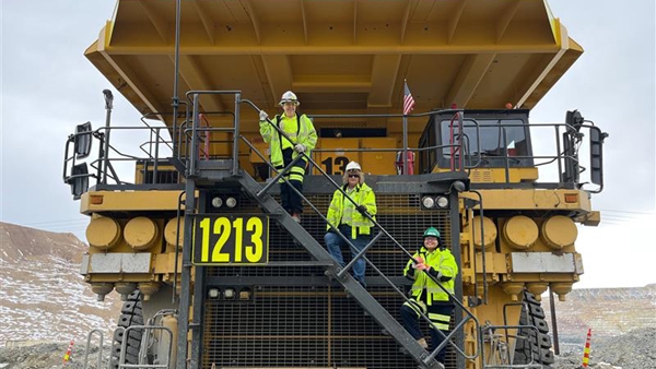 Three female workers wearing a high-visibility safety vest and helmet standing on stairs of a large haul truck.