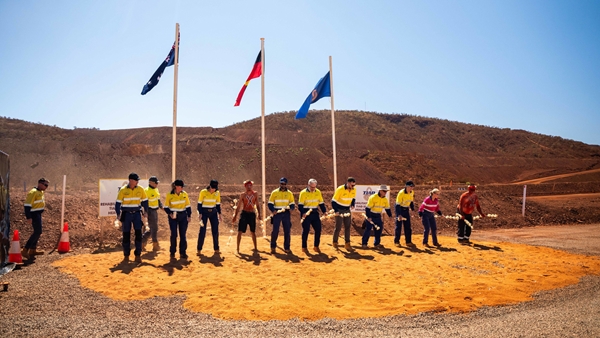 A group of approximately 13 people stand in a line on a large circular area of orange earth at a mining site, each holding handfuls of white flowers during a ceremonial event. Three flags fly on poles behind the group, with rugged red-brown hills and mine earthworks in the background under a clear blue sky. Several participants wear high-visibility yellow work shirts, while others wear different attire, highlighting a gathering that brings together mining personnel and community representatives in an outdoor industrial landscape.