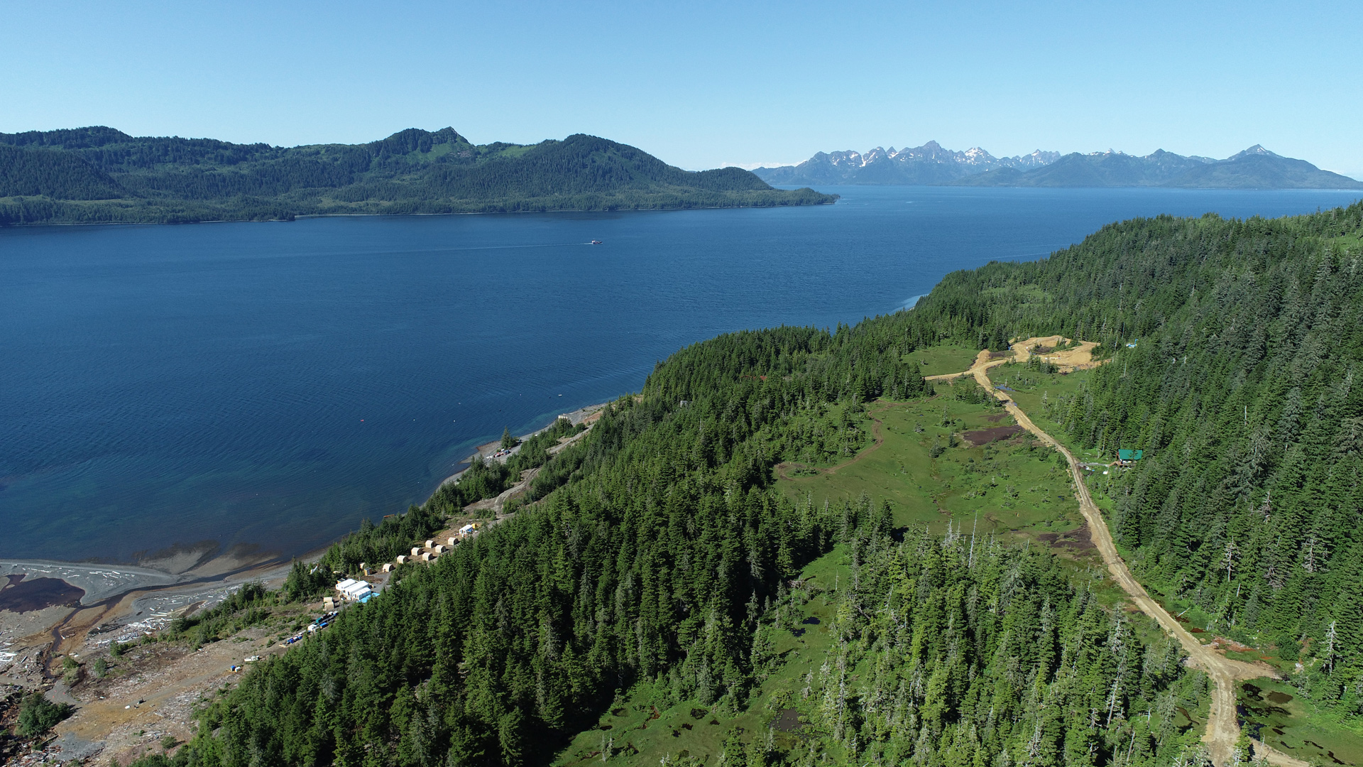 Aerial view of a forested peninsula beside a wide blue bay, with mountains rising in the distance.