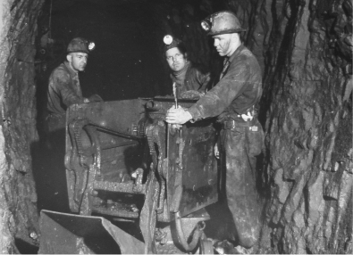 Black-and-white photograph of three people wearing hard hats with lamps, standing in a narrow underground tunnel and handling a metal cart filled with rock or ore, with rough rock walls surrounding them.