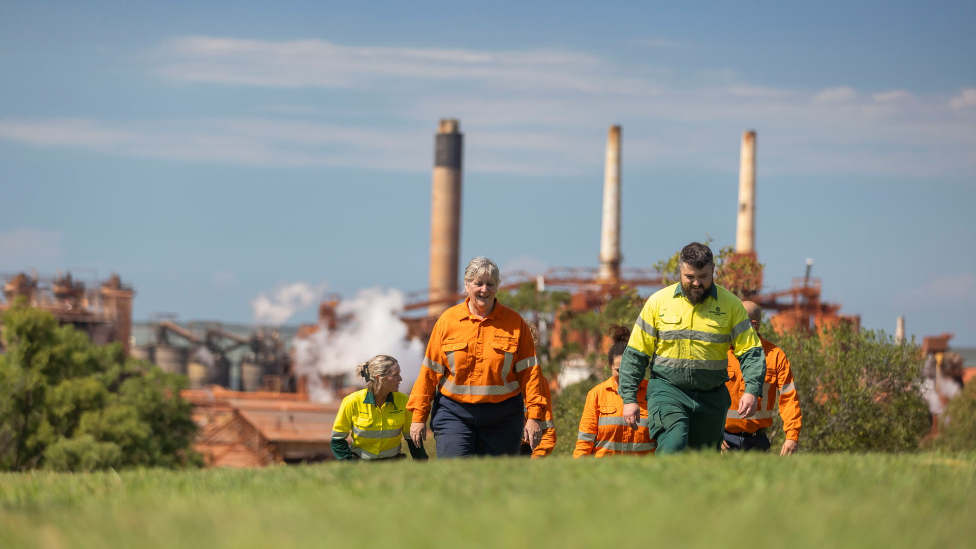 Industrial refinery viewed from a grassy area where workers in high-visibility clothing walking in the foreground.