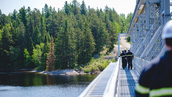 Un groupe de travailleurs portant des équipements de sécurité marche le long d'une structure métallique au-dessus d'un plan d'eau, avec une forêt dense en arrière-plan. Le pont est muni de garde-corps et semble faire partie d'une installation industrielle. Un autre travailleur est visible au premier plan, partiellement flou.