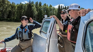 Trois personnes portant des gilets de sauvetage et des casquettes se tiennent côte à côte sur un petit bateau métallique, parlant près du poste de pilotage sur une large rivière boueuse. Des arbres bordent la rive en arrière-plan sous un ciel bleu dégagé, et la lumière du soleil éclaire la scène.
