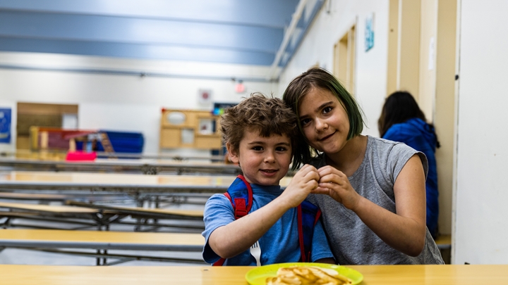 Deux jeunes enfants sont assis côte à côte à une table dans une cantine scolaire, tenant ensemble un petit objet et faisant face à la caméra. L’un des enfants porte une chemise bleue avec des bretelles de sac à dos rouges, et l’autre a les cheveux courts avec des mèches vertes et porte une chemise grise. Une assiette avec de la nourriture est visible au premier plan, tandis que les tables de la cantine et une autre personne assise à l’arrière-plan apparaissent légèrement floues.