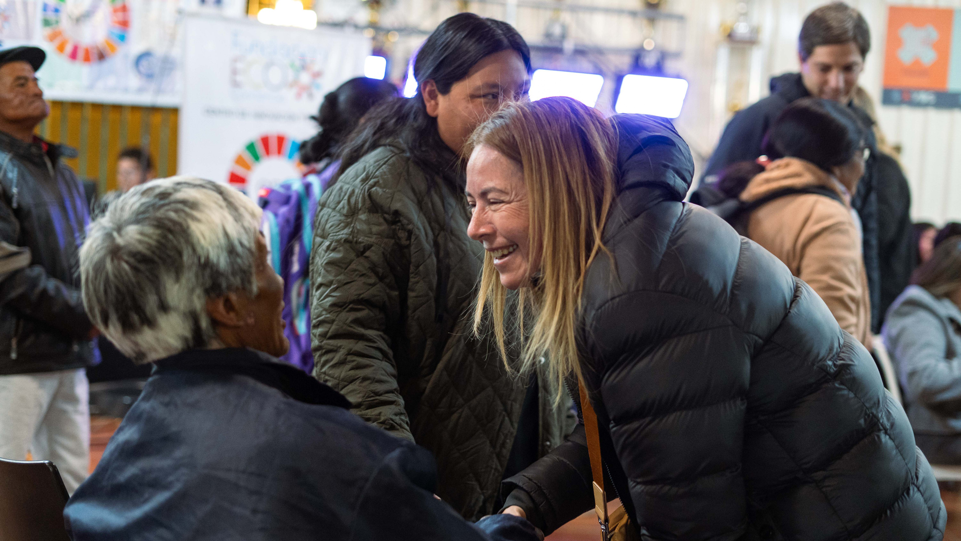 People greet and converse at a community gathering indoors, with participants seated and standing among informational displays and event signage in the background.