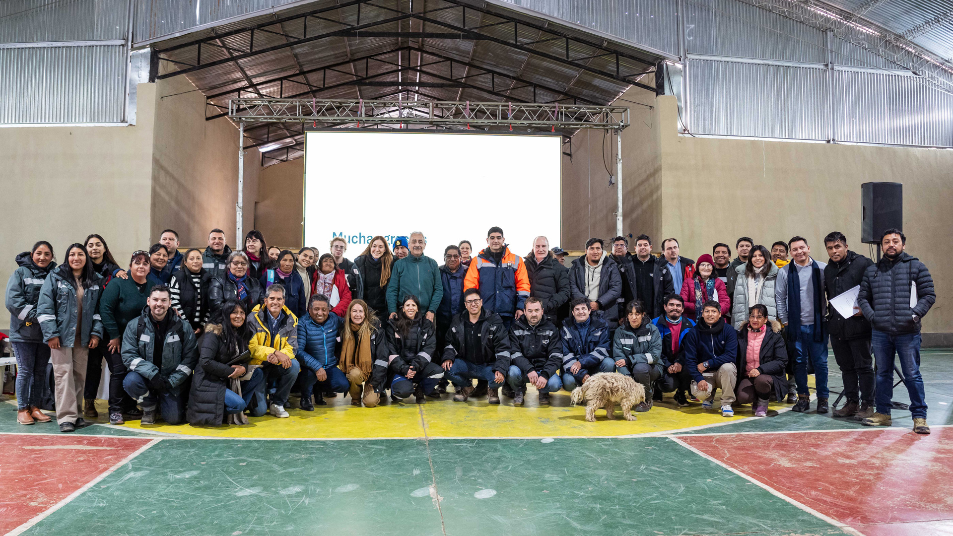 Large group photo of approximately 40 people gathered inside a covered indoor sports hall, posing on a basketball court in front of a projection screen and stage structure. Many participants are wearing work jackets and outdoor clothing, and a dog stands near the front row.