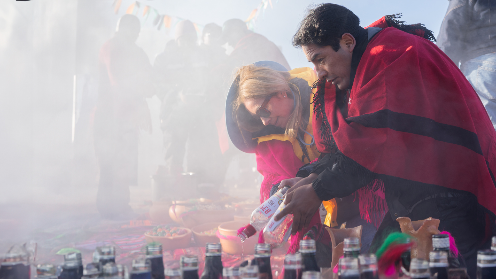 Two people wearing colorful shawls participate in a ceremonial gathering, pouring liquid into bowls amid a smoky outdoor setting.