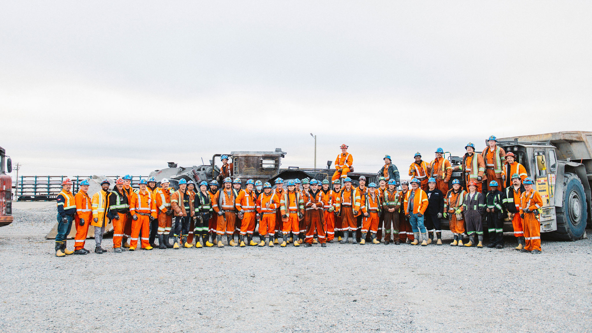 Large group of workers in high-visibility orange and reflective safety gear standing together on a gravel surface at an industrial site, with heavy mining trucks and equipment behind them under an overcast sky.